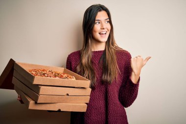 Young beautiful girl holding delivery boxes with Italian pizza standing over white background pointing and showing with thumb up to the side with happy face smiling