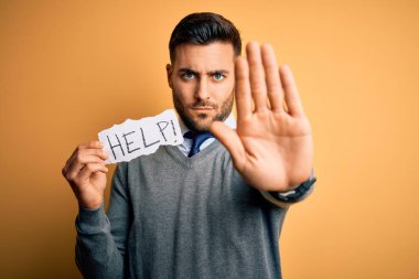 Young overworked business man asking for help holding paper over yellow background with open hand doing stop sign with serious and confident expression, defense gesture