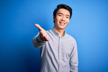 Young handsome chinese man wearing casual shirt standing over isolated blue background smiling friendly offering handshake as greeting and welcoming. Successful business.