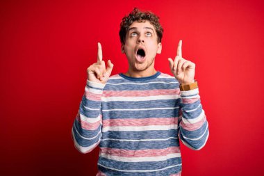 Young blond handsome man with curly hair wearing striped sweater over red background amazed and surprised looking up and pointing with fingers and raised arms.