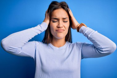 Young beautiful brunette woman wearing casual sweater standing over blue background suffering from headache desperate and stressed because pain and migraine. Hands on head.