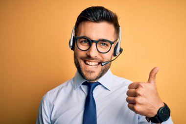 Young business operator man wearing customer service headset from call center doing happy thumbs up gesture with hand. Approving expression looking at the camera showing success.