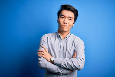 Young handsome chinese man wearing casual shirt standing over isolated blue background skeptic and nervous, disapproving expression on face with crossed arms. Negative person.