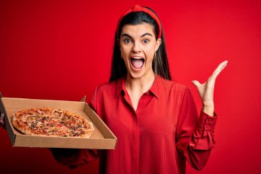 Genç, güzel esmer kadın elinde İtalyan pizzasıyla kırmızı arka planda mutlu ve heyecanlı bir şekilde teslimat kutusunu tutuyordu. Kazanan ifade zaferi büyük bir gülümsemeyle kutluyor ve ellerini kaldırıyordu.