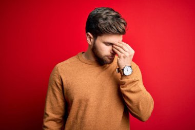 Young blond man with beard and blue eyes wearing casual sweater over red background tired rubbing nose and eyes feeling fatigue and headache. Stress and frustration concept.