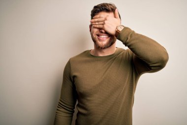 Young blond man with beard and blue eyes wearing green sweater over white background smiling and laughing with hand on face covering eyes for surprise. Blind concept.