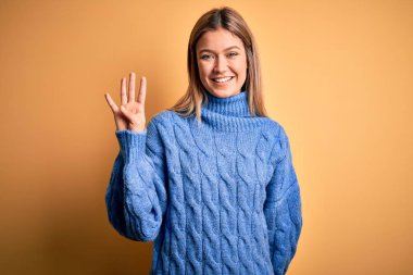 Sarıya boyanmış balıkçı yaka kazak giyen genç ve güzel bir kadın. İzole edilmiş sarı arka plan. Dört numaralı parmaklarıyla işaret ediyor..