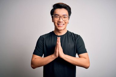 Young handsome chinese man wearing black t-shirt and glasses over white background praying with hands together asking for forgiveness smiling confident.