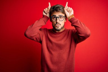 Young handsome man with beard wearing glasses and sweater standing over red background doing funny gesture with finger over head as bull horns