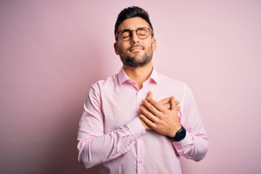 Young handsome man wearing elegant shirt and glasses standing over pink background smiling with hands on chest with closed eyes and grateful gesture on face. Health concept.