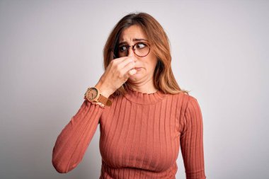 Young beautiful brunette woman wearing casual sweater and glasses over white background smelling something stinky and disgusting, intolerable smell, holding breath with fingers on nose. Bad smell