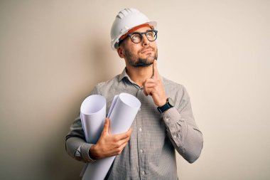 Young architect man wearing contractor helmet holding project paper plan over isolated background serious face thinking about question, very confused idea