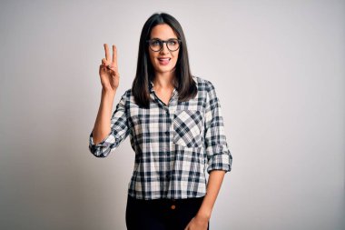 Mavi gözlü, sıradan gömlekli, beyaz arka planda gözlüklü, gülümseyen ve parmaklarını zafer işareti yaparken gösteren genç esmer kadın. İki numara..