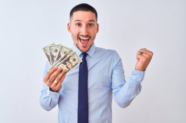 Young handsome business man holding a bunch of bank notes dollars over isolated background screaming proud and celebrating victory and success very excited, cheering emotion