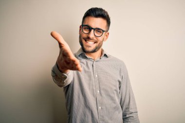 Young handsome man wearing elegant shirt and glasses over isolated white background smiling friendly offering handshake as greeting and welcoming. Successful business.