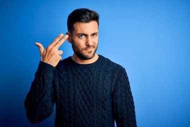 Young handsome man wearing casual sweater standing over isolated blue background Shooting and killing oneself pointing hand and fingers to head like gun, suicide gesture.