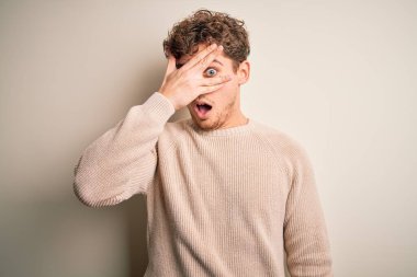 Young blond handsome man with curly hair wearing casual sweater over white background peeking in shock covering face and eyes with hand, looking through fingers with embarrassed expression.