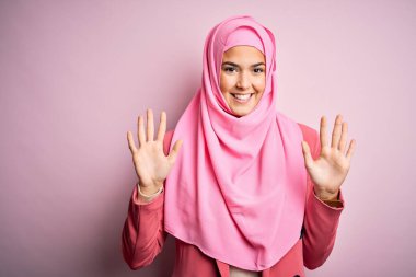 Young beautiful girl wearing muslim hijab standing over isolated pink background showing and pointing up with fingers number ten while smiling confident and happy.
