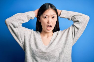 Genç ve güzel Asyalı bir kadın mavi izole edilmiş arka planda dikilen günlük süveter giyiyor. Deli ve elleri başında korkmuş, korkmuş ve ağzı açık bir şekilde şok geçirmekten korkan.