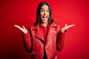 Genç ve güzel esmer bir kadın, gündelik ceketini giymiş, kırmızı arka planda durmuş çılgın ve başarıyı kutluyor, kollarını kaldırmış ve gözleri açık, heyecanlı bir şekilde çığlık atıyor. Kazanan konsept