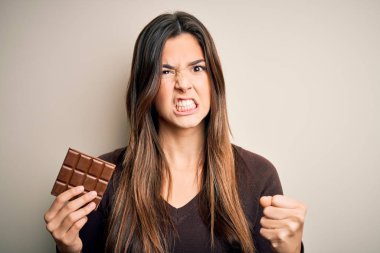 Young beautiful girl holding sweet bar of chocolate over isolated white background annoyed and frustrated shouting with anger, crazy and yelling with raised hand, anger concept