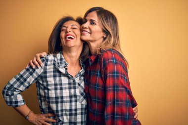 Orta yaşlı, güzel iki kız kardeş mutlu ve kendinden emin gülümsüyor. Yüzünde gülümsemeyle dikilip izole edilmiş sarı arka planda sarılıyorum.