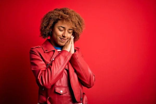 Young beautiful African American afro woman with curly hair wearing casual red jacket sleeping tired dreaming and posing with hands together while smiling with closed eyes.