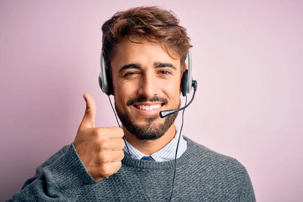 Young call center agent man with beard wearing headset over isolated pink background doing happy thumbs up gesture with hand. Approving expression looking at the camera showing success.