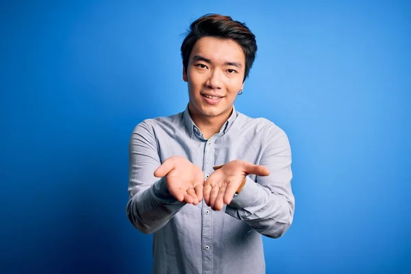 Young handsome chinese man wearing casual shirt standing over isolated blue background Smiling with hands palms together receiving or giving gesture. Hold and protection