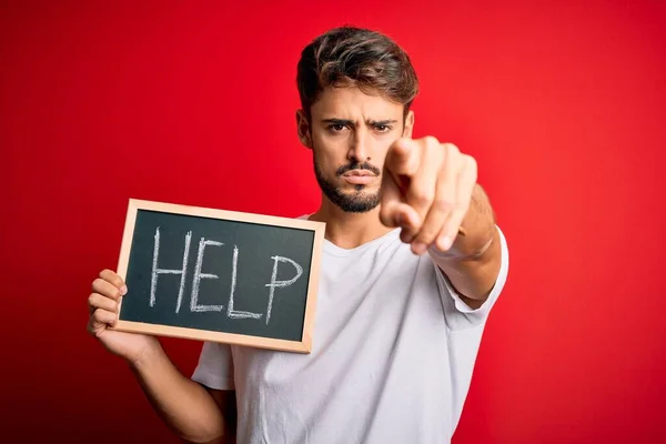 Young man with problem holding blackboard with help message over red bakground pointing with finger to the camera and to you, hand sign, positive and confident gesture from the front