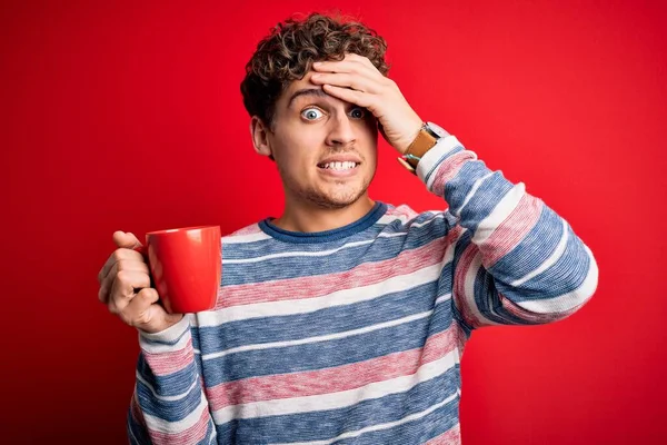 Young blond man with curly hair drinking cup of coffee standing over red background stressed with hand on head, shocked with shame and surprise face, angry and frustrated. Fear and upset for mistake.