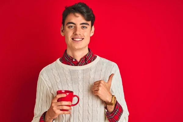 ティーネージャー男の子身に着けている冬のセーターを飲みますコーヒーのカップ以上隔離された赤の背景幸せなで大きな笑顔を行いますOkサイン 親指アップとともに指 優れたサイン — ストック写真