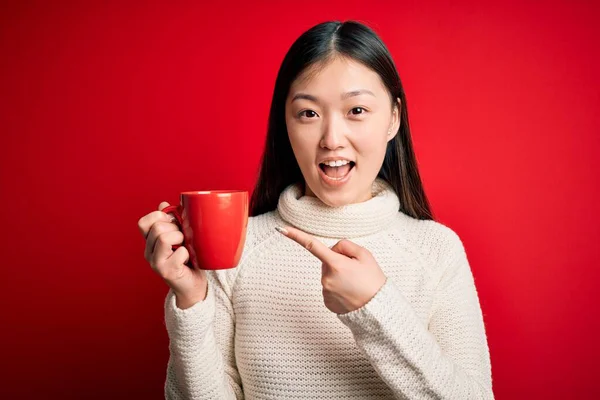年轻的亚洲女人在孤立的红色背景下喝着一杯热咖啡 她非常高兴地用手和手指指指点点 — 图库照片