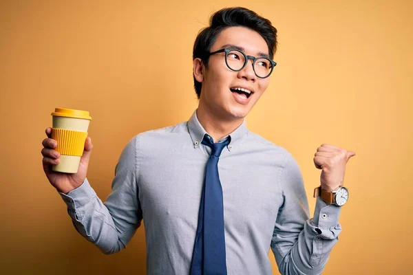 Young handsome chinese man drinking glass of take away coffee over yellow background pointing and showing with thumb up to the side with happy face smiling