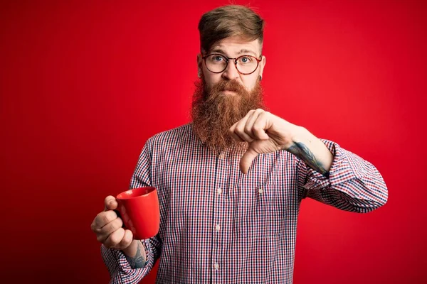Redhead Irish man with beard drinking a cup of hot coffee over red background with angry face, negative sign showing dislike with thumbs down, rejection concept