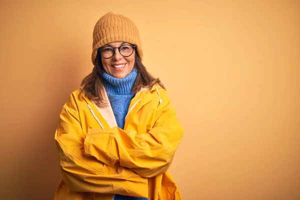 Middle age woman wearing yellow raincoat and winter hat over isolated background happy face smiling with crossed arms looking at the camera. Positive person.