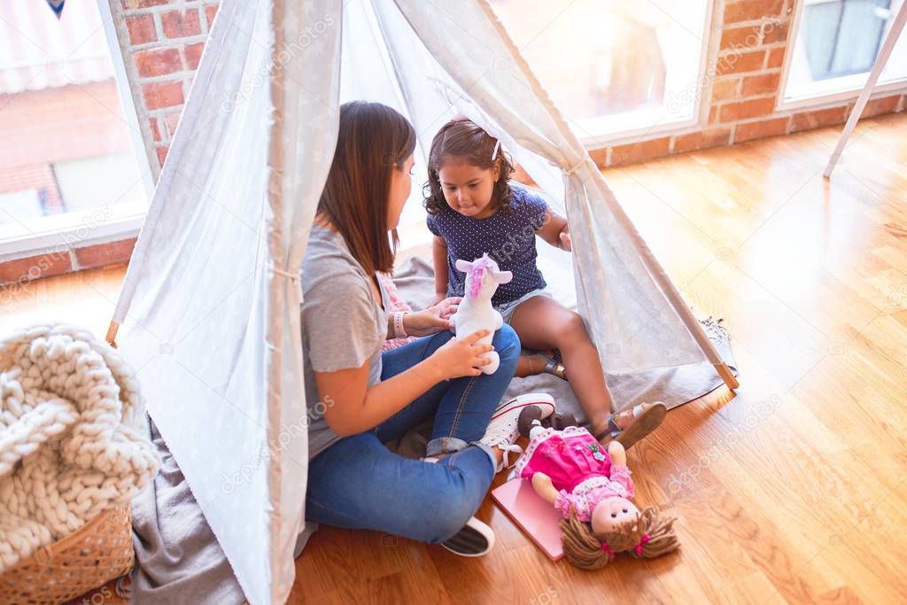 Beautiful teacher and toddler girl sitting on the floor playing with unicorn and doll inside tipi at kindergarten