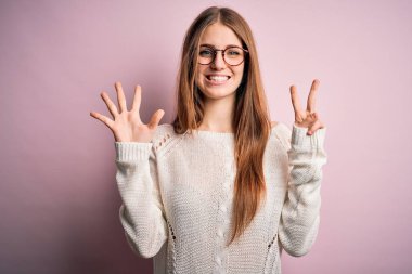 Young beautiful redhead woman wearing casual sweater and glasses over pink background showing and pointing up with fingers number seven while smiling confident and happy.
