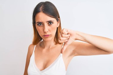 Güzel kızıl saçlı kadın, izole edilmiş arka planda günlük beyaz tişört giyiyor. Kızgın yüzlü, negatif işaretli, baş parmakları aşağıda, reddedilme kavramından hoşlanmadığını gösteriyor.