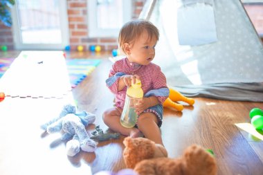 Anaokulunda bir sürü oyuncağın etrafında beslenme şişesi tutan sevimli çocuk.