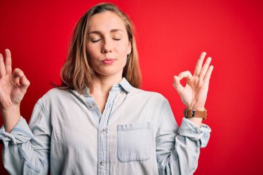 İzole edilmiş kırmızı arka plan üzerinde sıradan bir kot gömlek giyen genç, güzel sarışın kadın rahatla ve parmaklarıyla meditasyon hareketi yaparken gözleri kapalı gülümse. Yoga konsepti.