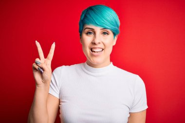 Mavi saçlı, kırmızı arka planda rahat tişörtler giyen genç ve güzel bir kadın iki numarayı gösterirken kendinden emin ve mutlu bir şekilde gülümsüyor..