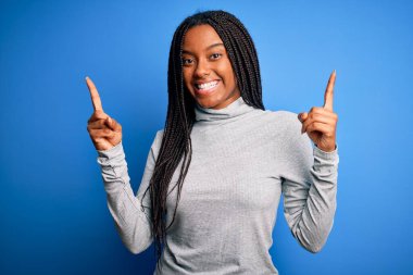 Genç Afrikalı Amerikalı kadın mavi izole edilmiş arka planda balıkçı yaka kazak giyiyor ve parmaklarıyla farklı yönleri işaret ederek gülümsüyor. Reklam için boşluk kopyala