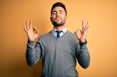 Zarif bir süveter giyen ve sarı arka planda kravat takan genç yakışıklı işadamı rahatla ve parmaklarıyla meditasyon yaparken gözleri kapalı gülümse. Yoga konsepti.
