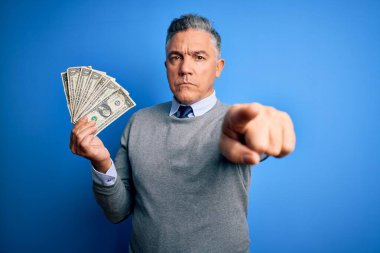 Orta yaşlı, yakışıklı, gri saçlı, elinde bir tomar parayla kamerayı işaret eden bir adam ve sen, el işareti, ön taraftan pozitif ve kendinden emin bir jest.