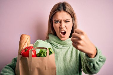 Genç, sarışın bir kız pembe izole edilmiş arka planda yeni paketlenmiş paketler taşıyor. Öfkeyle bağırıyor, sinirleniyor ve sinirleniyor. Öfkeyle bağırıyor.