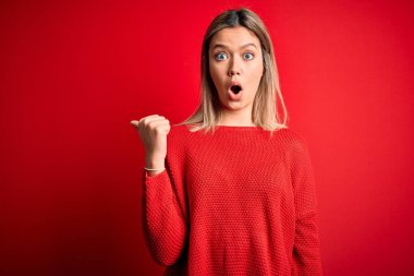 Kırmızı izole edilmiş arka plan üzerine gündelik süveter giyen genç, güzel sarışın bir kadın. El parmağı yana dönük, ağzı açık, şaşkın bir ifade..