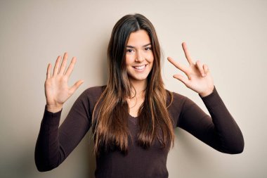 İzole edilmiş beyaz arka plan üzerinde duran sıradan süveter giyen genç ve güzel bir kız sekiz numaralı parmaklarıyla işaret ederken kendinden emin ve mutlu bir şekilde gülümsüyor..