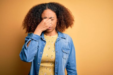 Afro saçlı, sarı arka plan üzerine günlük kot gömlek giyen, burnu ve gözleri ovuşturan yorgun, yorgun ve başı ağrıyan Afro-Amerikan genç bir kadın. Stres ve hüsran kavramı.