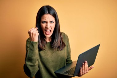 Mavi gözlü genç esmer kadın bilgisayarını kullanarak sarı arka plan üzerinde çalışıyor kızgın ve kızgın yumruğunu kaldırıyor ve öfkeyle bağırıyor. Öfke ve agresif kavram.